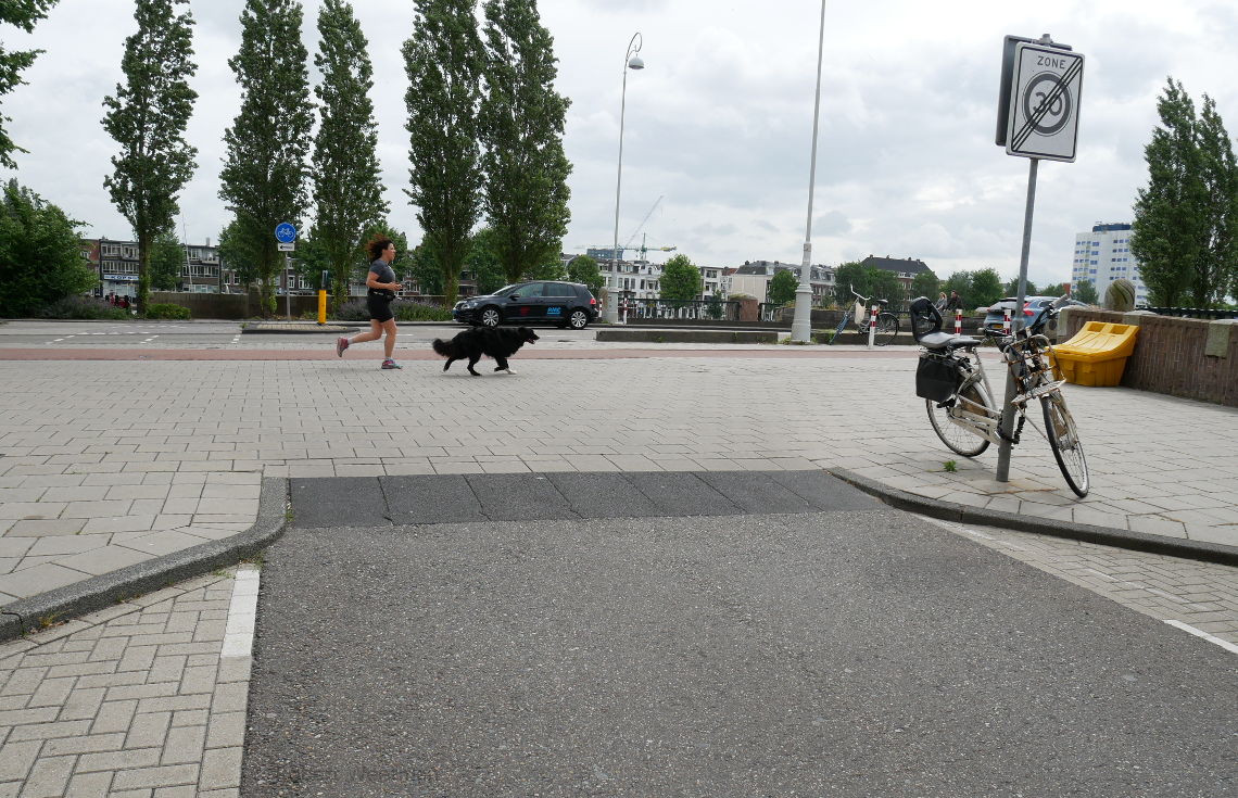 Image shows a woman and dog running along a piece of footway over which people might drive to access a side road.