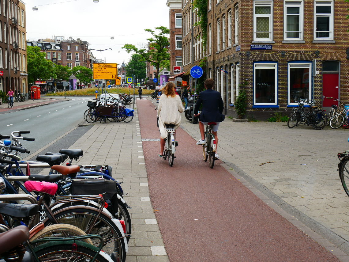 Image shows two people cycling along a cycle track talking to one another, paying little attention to the roads to either side of them, as the track passes across a section of continuous footway.